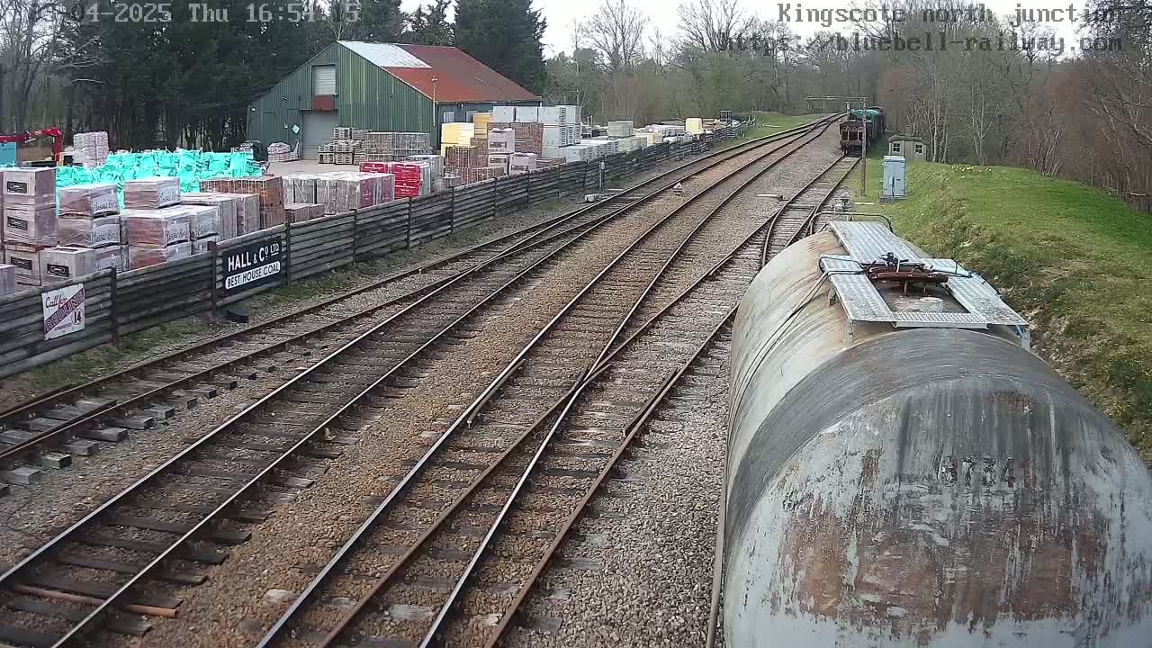A railway junction on a partly sunny day, showing several railway tracks, a building with stacked building materials beside it, and a train car in the foreground.