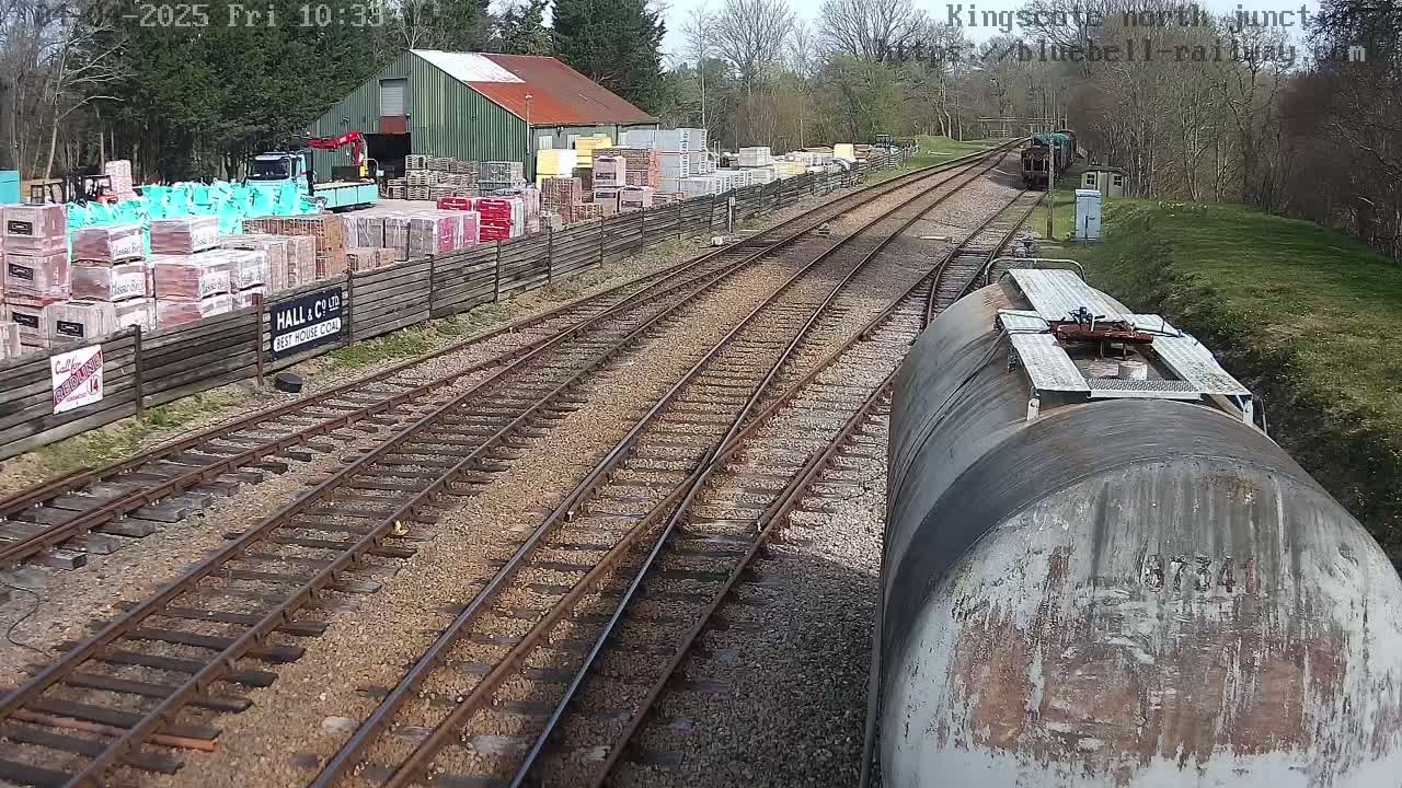 A view from a train shows a railway junction with multiple tracks, a building materials yard beside it, and a train car in the foreground; the weather is sunny.