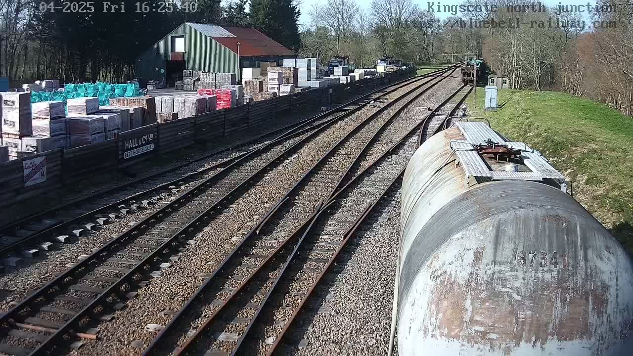 A railway junction on a sunny day, viewed from a train, showing multiple railway tracks branching off from a main line, with a building and stacks of building materials nearby.
