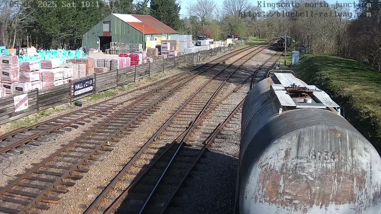A view from a train car shows a sunny day at a railway junction with a building and stacks of building materials alongside the tracks.
