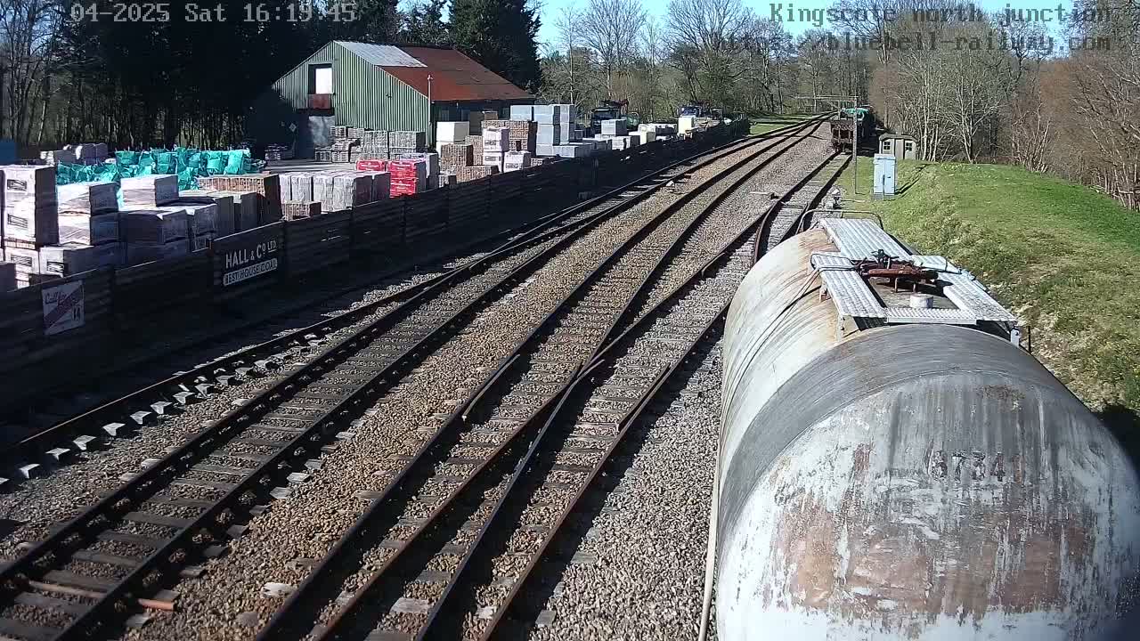 A sunny day at a railway junction shows several train tracks converging near a building with stacks of building materials, and a weathered tanker car is in the foreground.