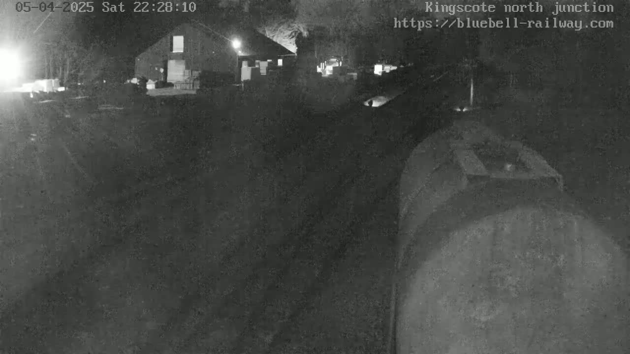 A nighttime, low-light view of a dark, seemingly industrial area with a building in the background, and a large, dark object in the foreground that resembles a piece of heavy machinery or a vehicle.
