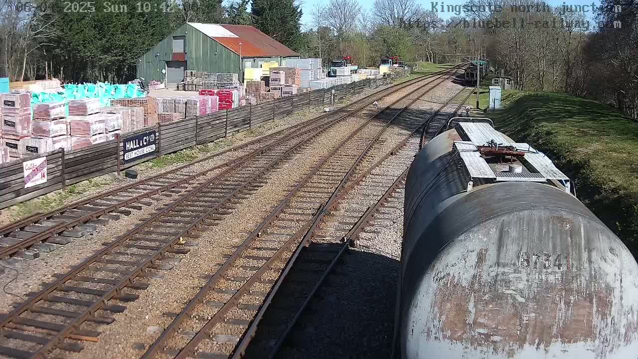 A view from a train car shows railway tracks leading to a building and yard stocked with bricks and other building materials, under a sunny sky.