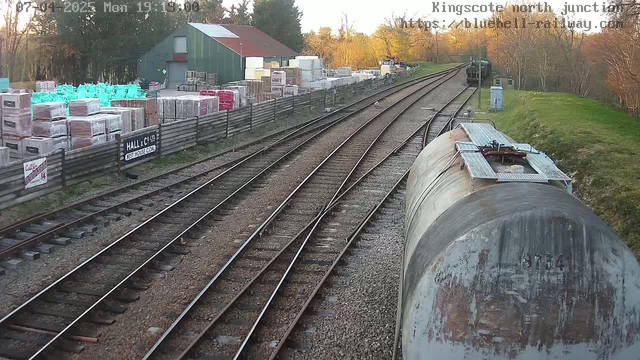 A view from a train car shows a railway junction with multiple tracks, a building containing stacks of building materials, and a partly sunny day.