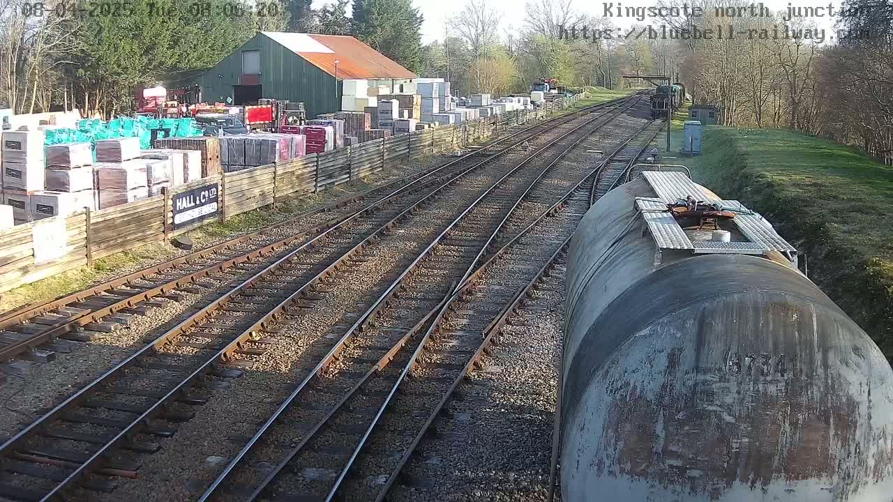 A view from a train shows multiple train tracks converging at a junction, with a building and stacks of materials visible to the side on a sunny day.