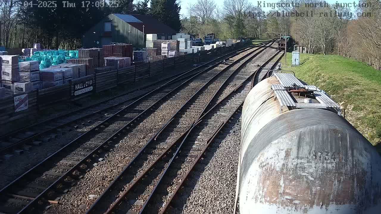 A train car sits on a railway track next to a building and a yard filled with stacked building materials on a sunny day.