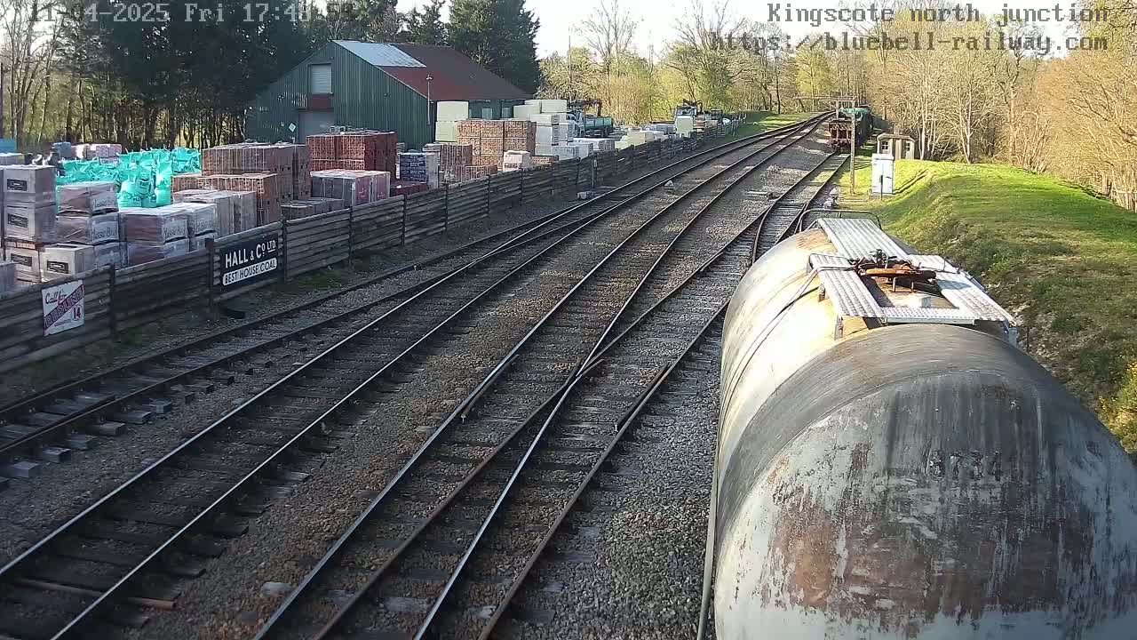 A view from a train of railway tracks branching out from a junction, passing alongside a building and stacks of building materials in a sunny outdoor setting.