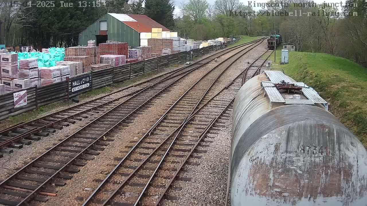 A view from a railway car shows train tracks branching off, with a building and stacks of building materials to the left, and a partly cloudy sky overhead.