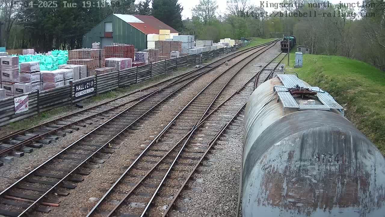A train on a railway track passes a building and a yard full of stacked building materials on an overcast day.