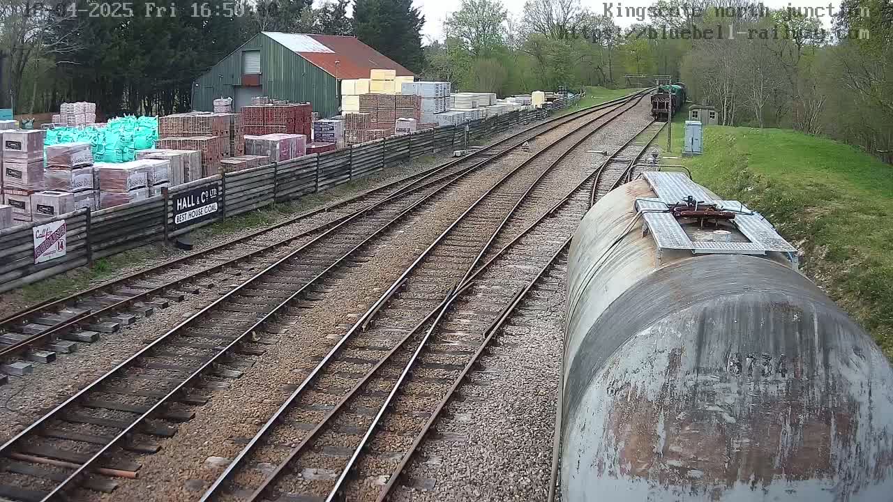 A railway yard with multiple tracks, a building containing stacked materials, and a train car in the foreground on a partly cloudy day.
