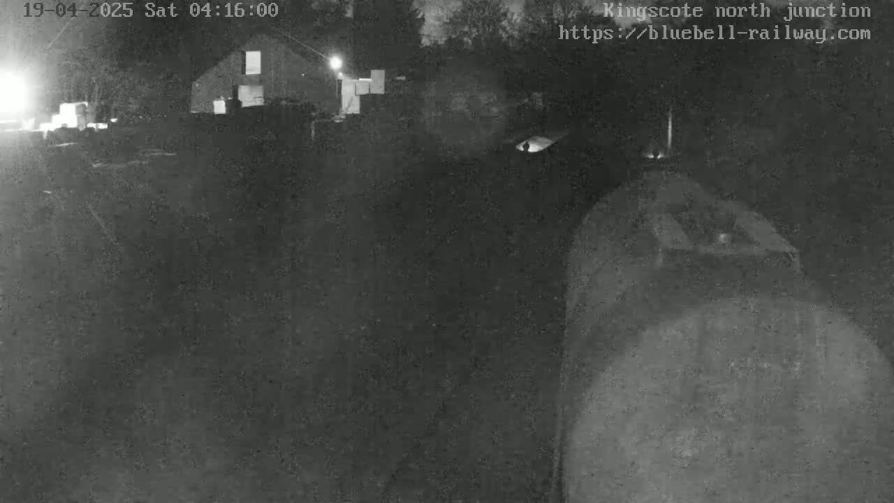 A nighttime, low-light image shows a dark, possibly industrial area with a building in the background and what appears to be a large piece of machinery or vehicle in the foreground.
