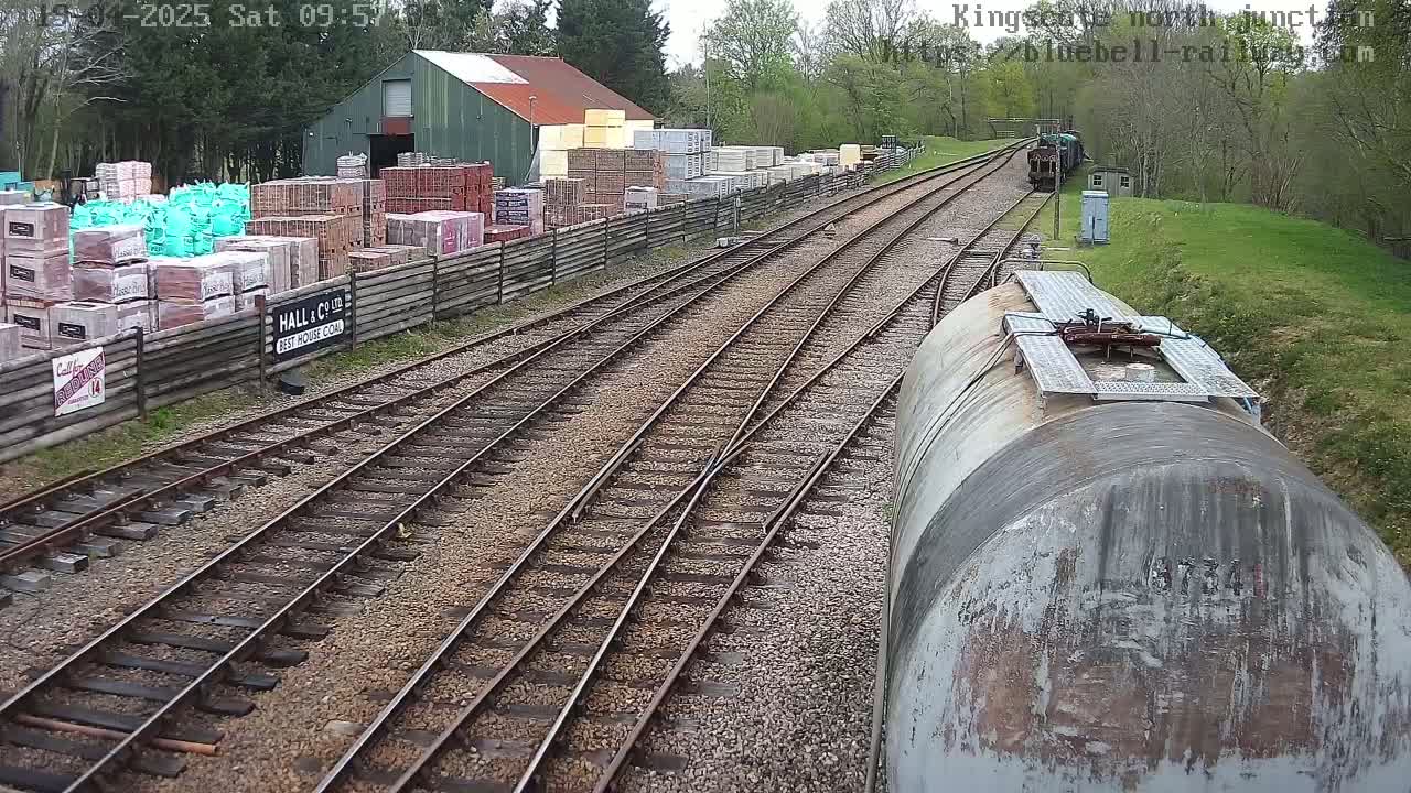 A view from a train car shows a railway junction, a building with stacked building materials alongside the tracks, and green trees in the background under a clear sky.