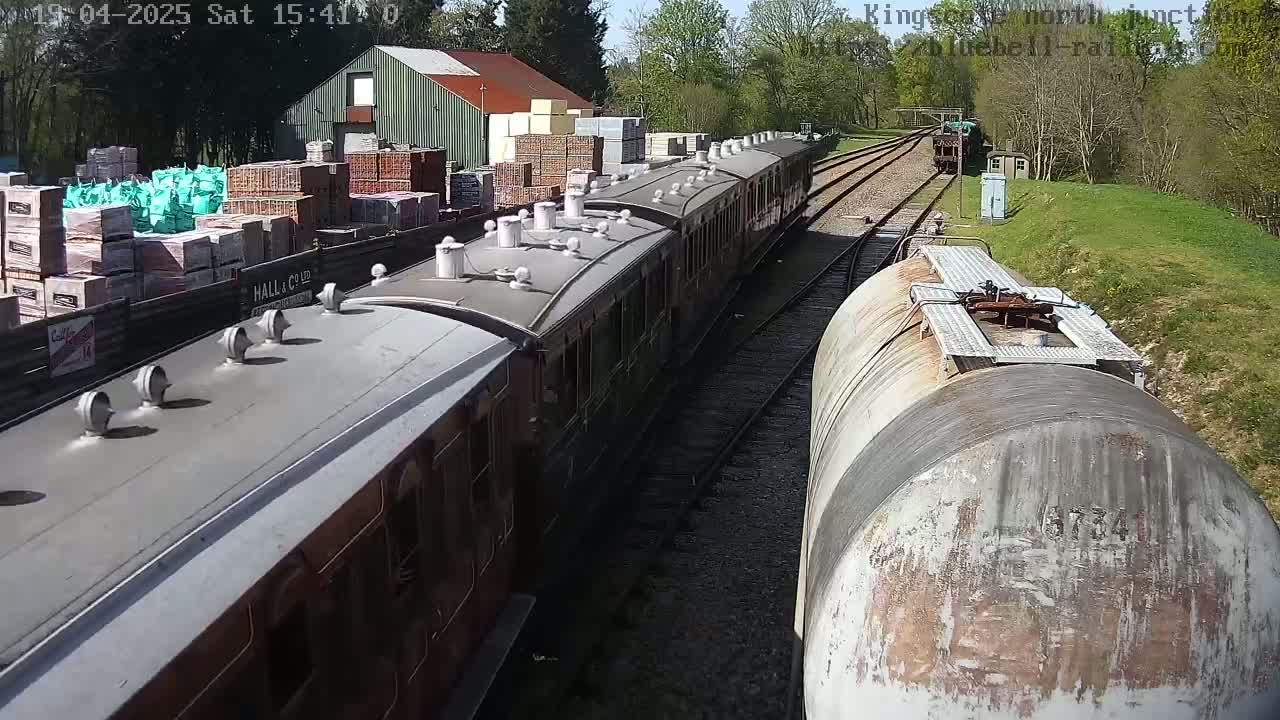 A passenger train and a tanker car are on a railway track next to a building and a pile of building materials on a sunny day.