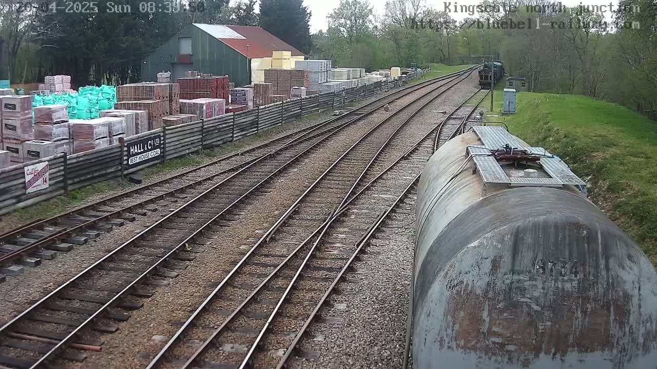 A train car sits on a railway track next to a building and a yard stacked with building materials, on a sunny day.