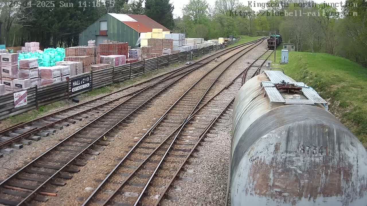 A view from a train car shows railway tracks branching off towards a building and yard with stacks of building materials under a partly cloudy sky.