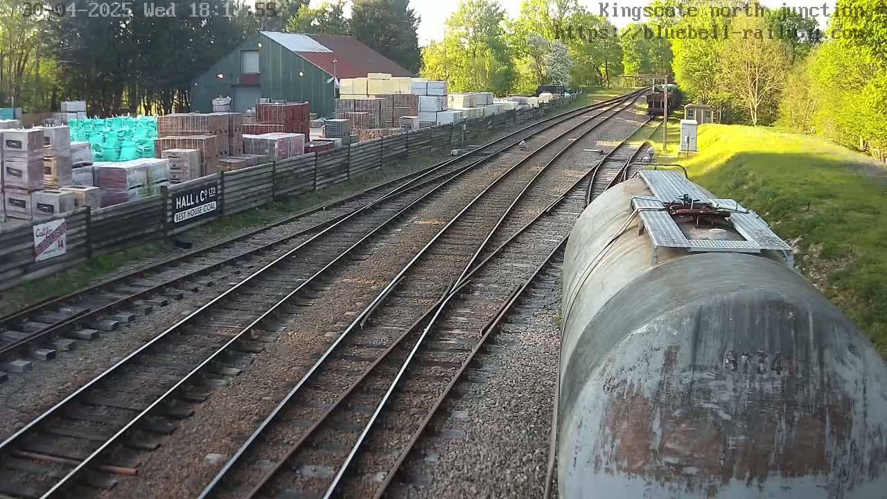 A view from a train shows multiple train tracks converging, alongside a building containing stacked building materials, under a partly sunny sky.