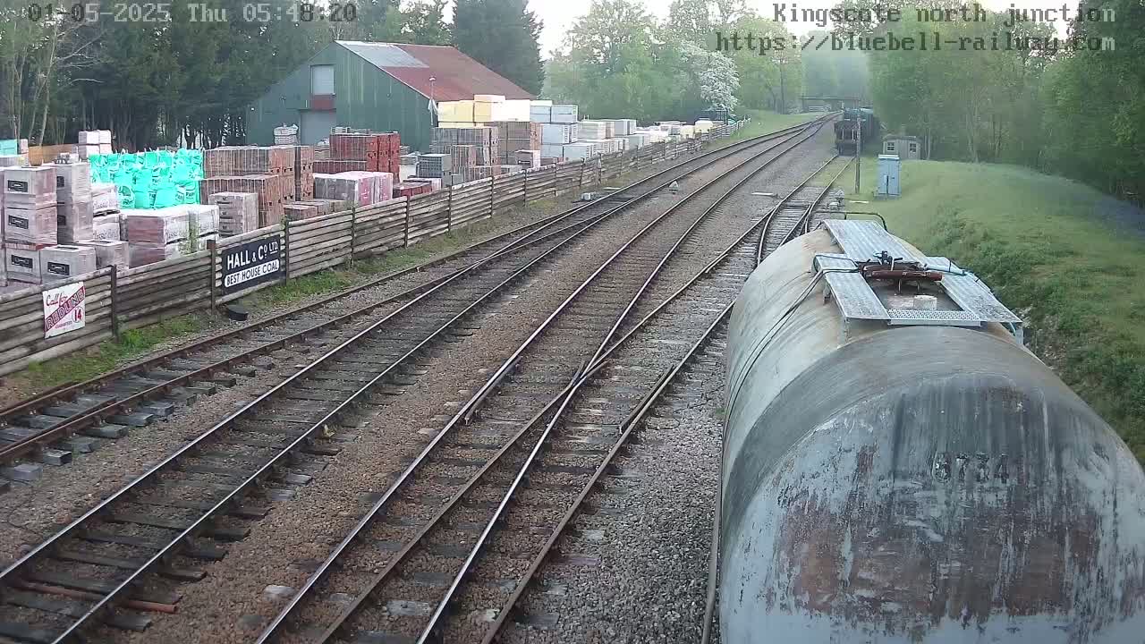 A railway junction with multiple tracks, a weathered rail tank car, and a building with stacks of building materials in the background under an overcast sky.