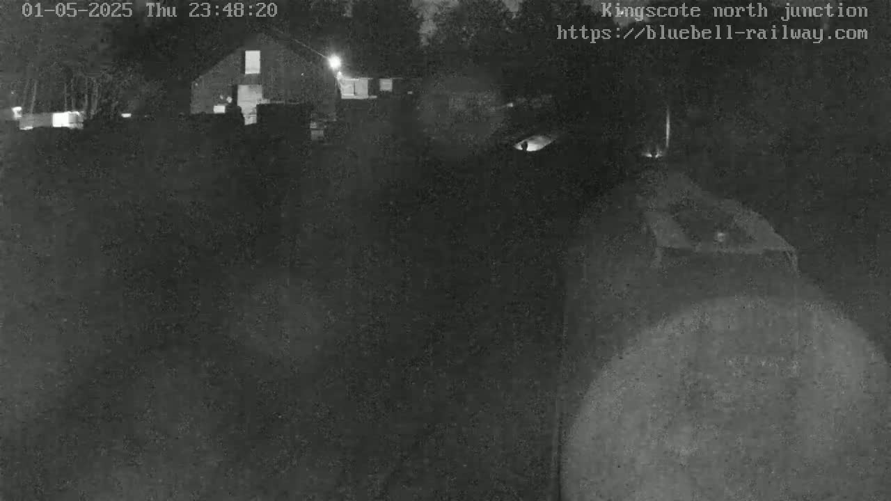 A nighttime, low-light image shows a dark, indistinct scene with a barn and what appears to be a vehicle in the foreground.