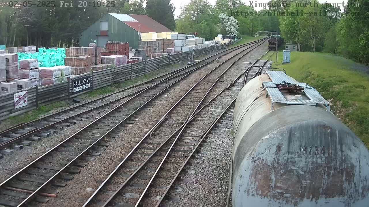 A view from a train car shows multiple train tracks converging, with a building and stacks of building materials to the left, and a wooded area in the background under a partly cloudy sky.
