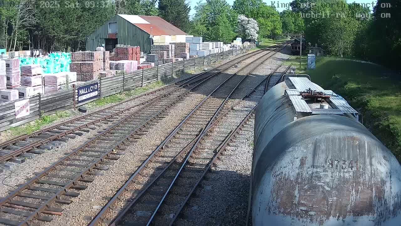 A view from a train of railway tracks splitting, leading to a building yard filled with stacks of building materials under a sunny sky.
