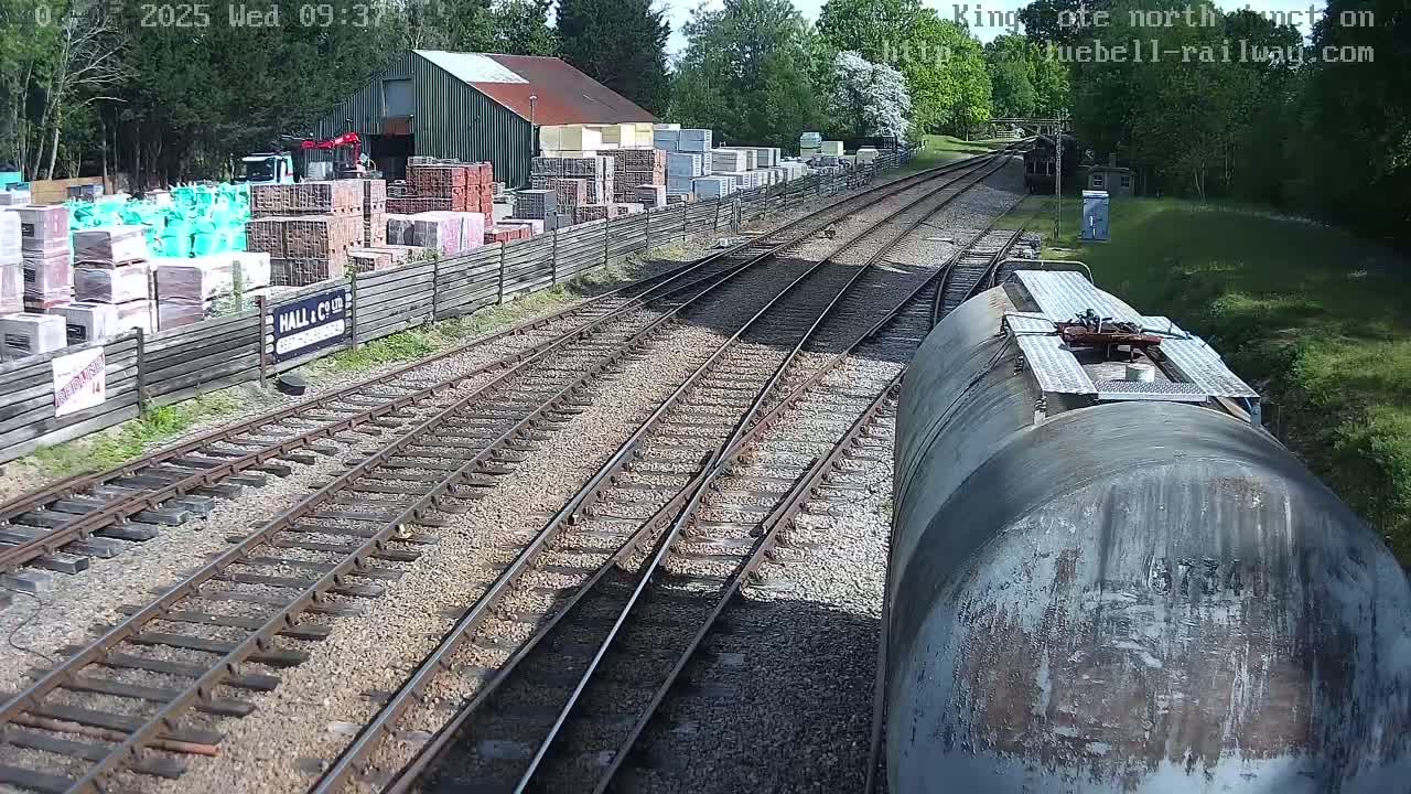 A view from a train car shows railway tracks splitting into two, alongside a building and stacks of building materials in the sunny outdoors.