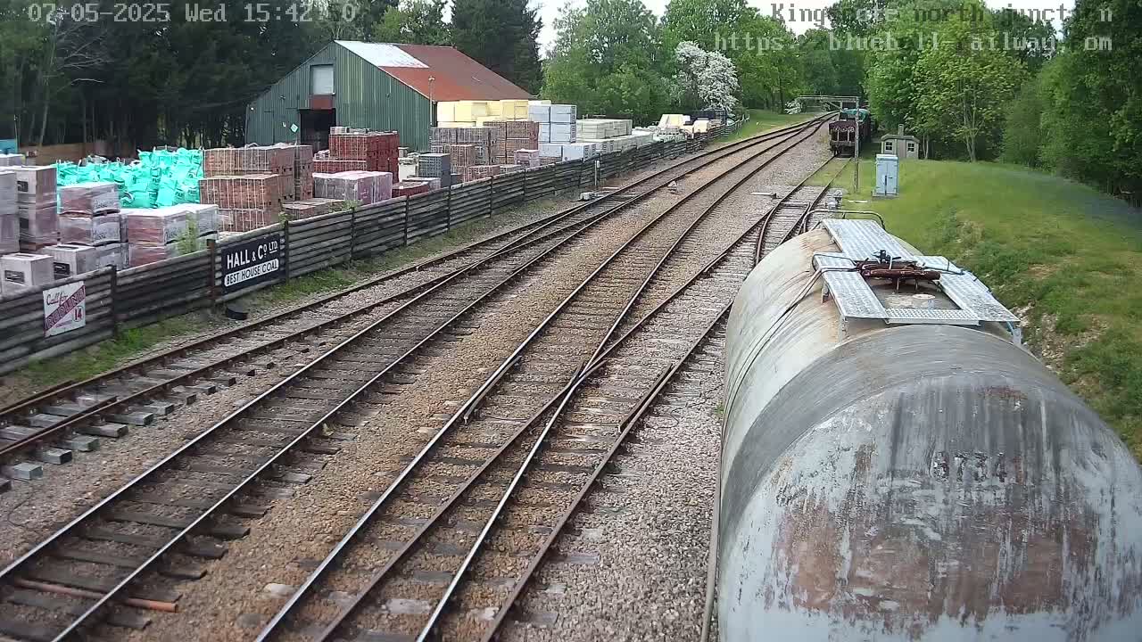 A view from a train shows several railway tracks branching off, alongside a building and stacks of building materials, in a setting with trees and under a partly cloudy sky.