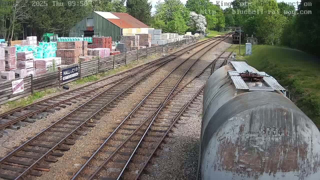 A view from a train car shows railway tracks branching out from a yard on a sunny day, alongside a building with stacks of bricks and other building materials.