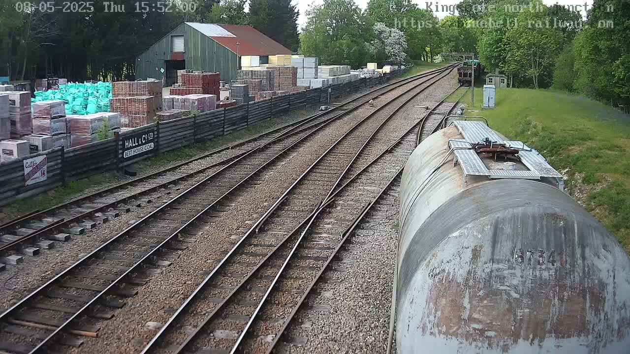 A view from a train of railway tracks branching off, beside a building and yard stacked with building materials, under a partly cloudy sky.