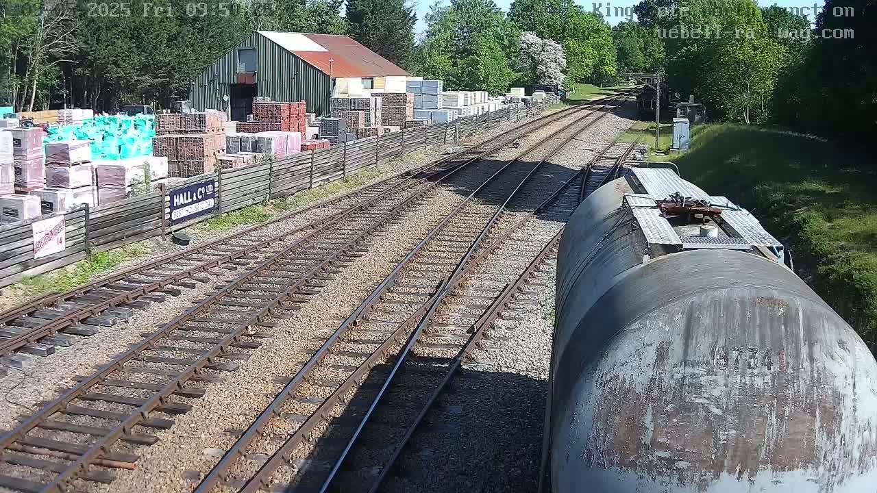 A view from a train car shows railway tracks branching out, passing by a building and a yard stacked with building materials, under a sunny sky.