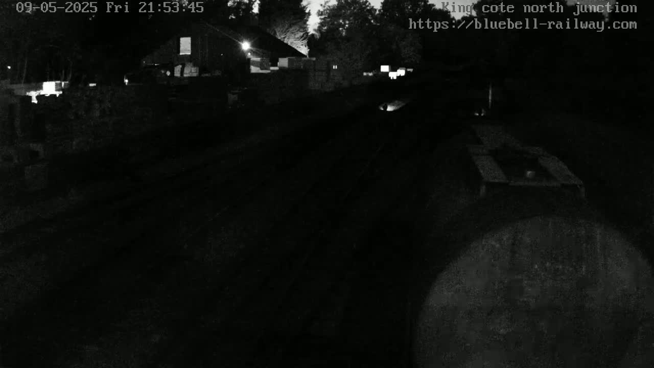 A nighttime, low-light view of a dark railway track leading toward a building and some stacked materials, under a mostly clear sky.