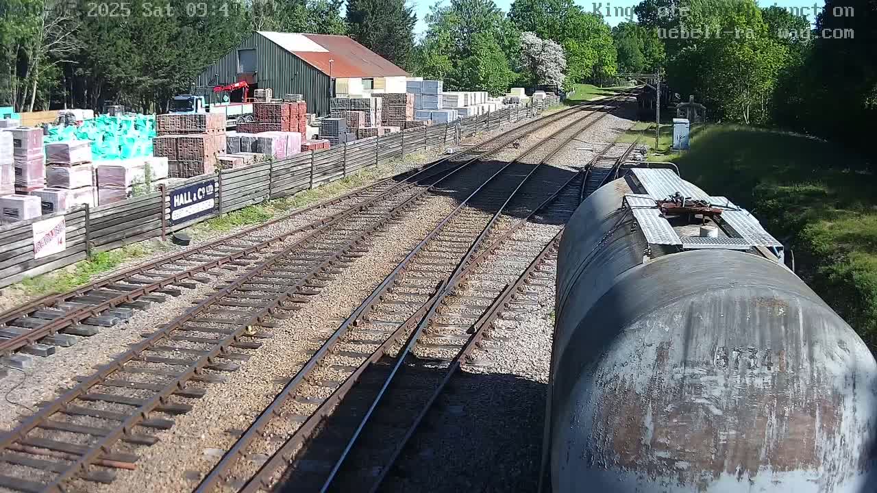 A view from a train shows railway tracks branching off near a building and a yard stacked with building materials, all under sunny conditions.