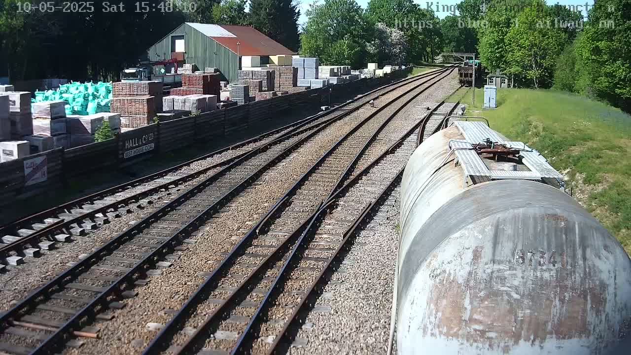 A view from a train car shows railway tracks branching out from a yard with building materials stacked beside a building, all under a sunny sky.