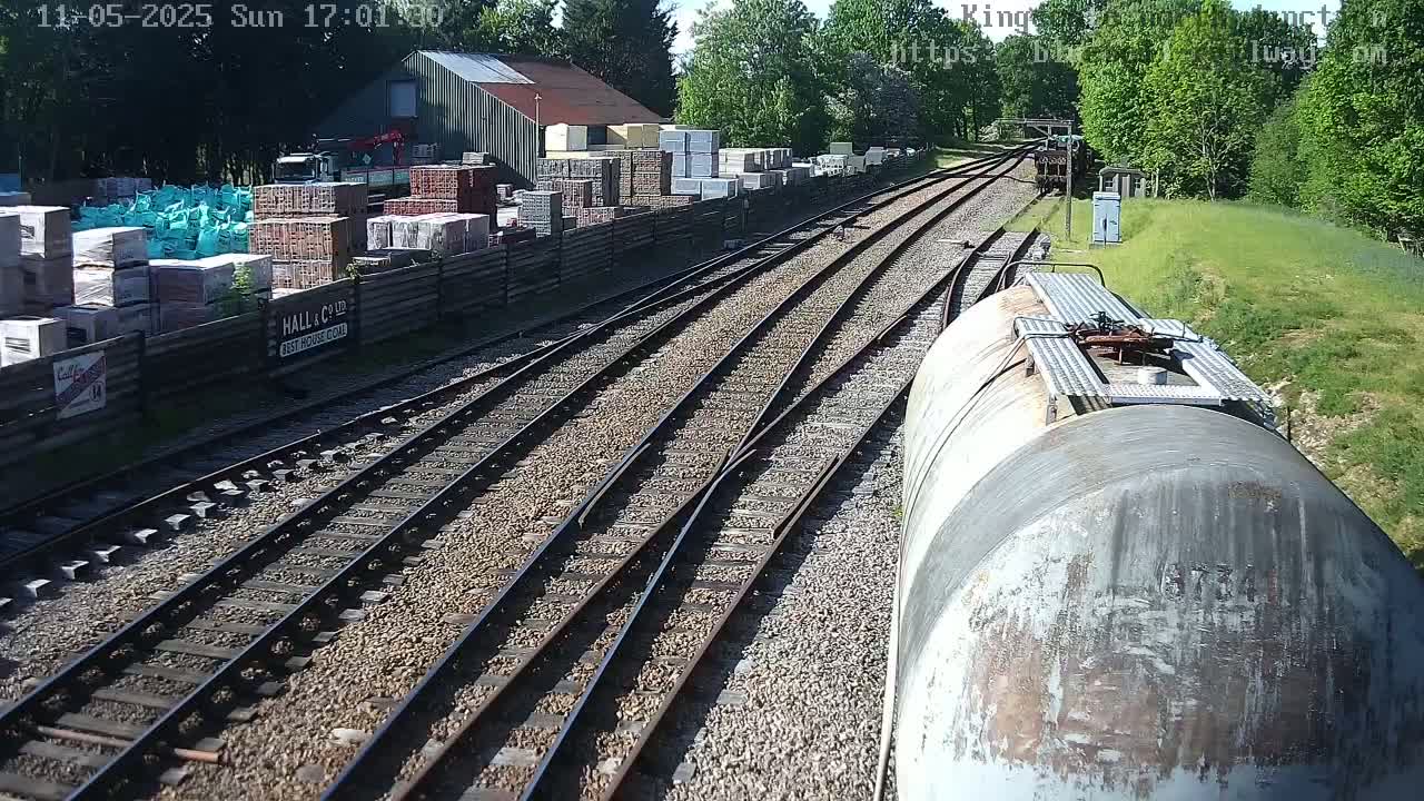 A view from a train car shows a sunny day at a railway yard with multiple train tracks branching out, alongside a building containing stacked building materials.