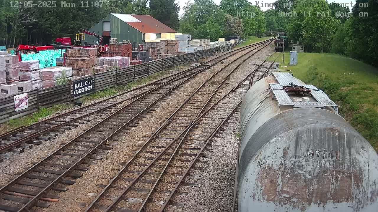 A railway yard with multiple tracks, a building storing building materials, and a weathered tanker car is seen on a partly sunny day.