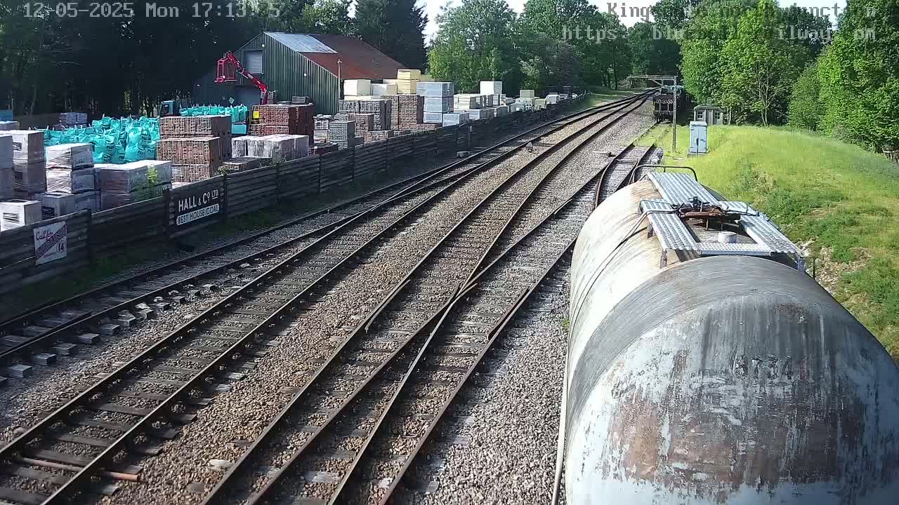 A train passes railway tracks next to a building and a yard full of stacked building materials on a sunny day.
