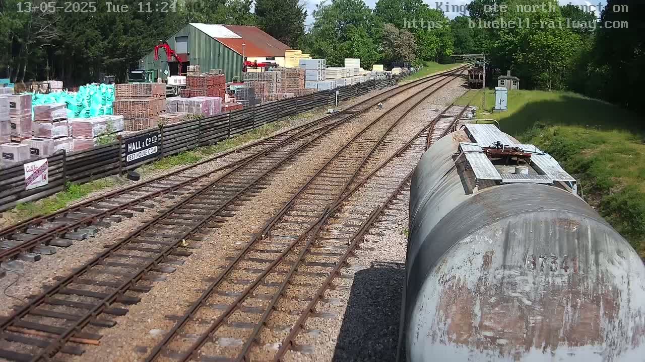 A railway track winds through a sunny landscape past a building materials yard and a weathered tank car.