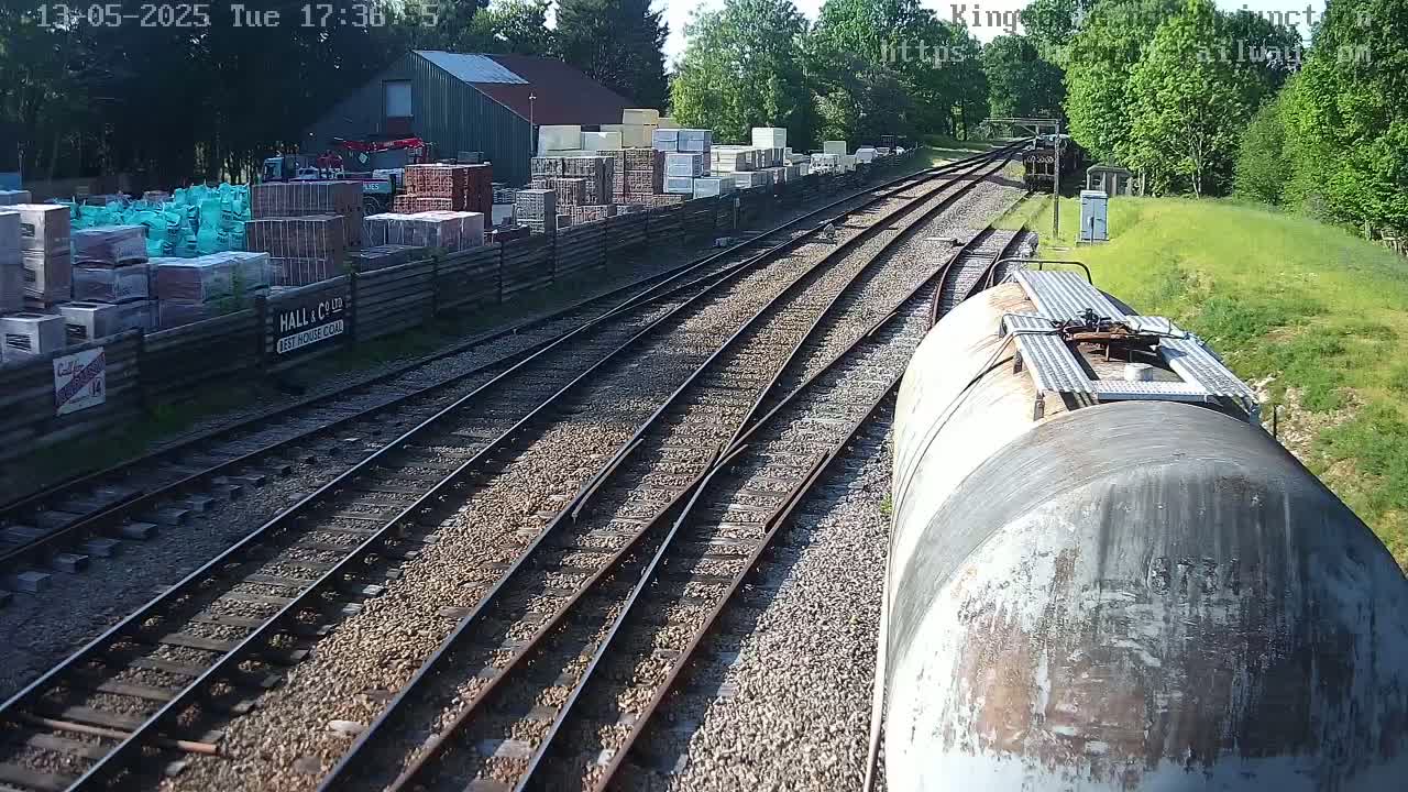 A view from a train car shows a sunny day at a railway yard with multiple train tracks converging, alongside a building and stacks of building materials.