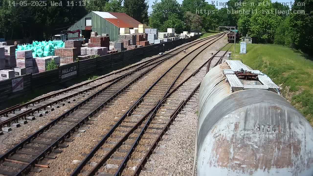 A view from a train car shows a sunny day overlooking train tracks splitting into two directions, with a building and stacks of building materials to the left.