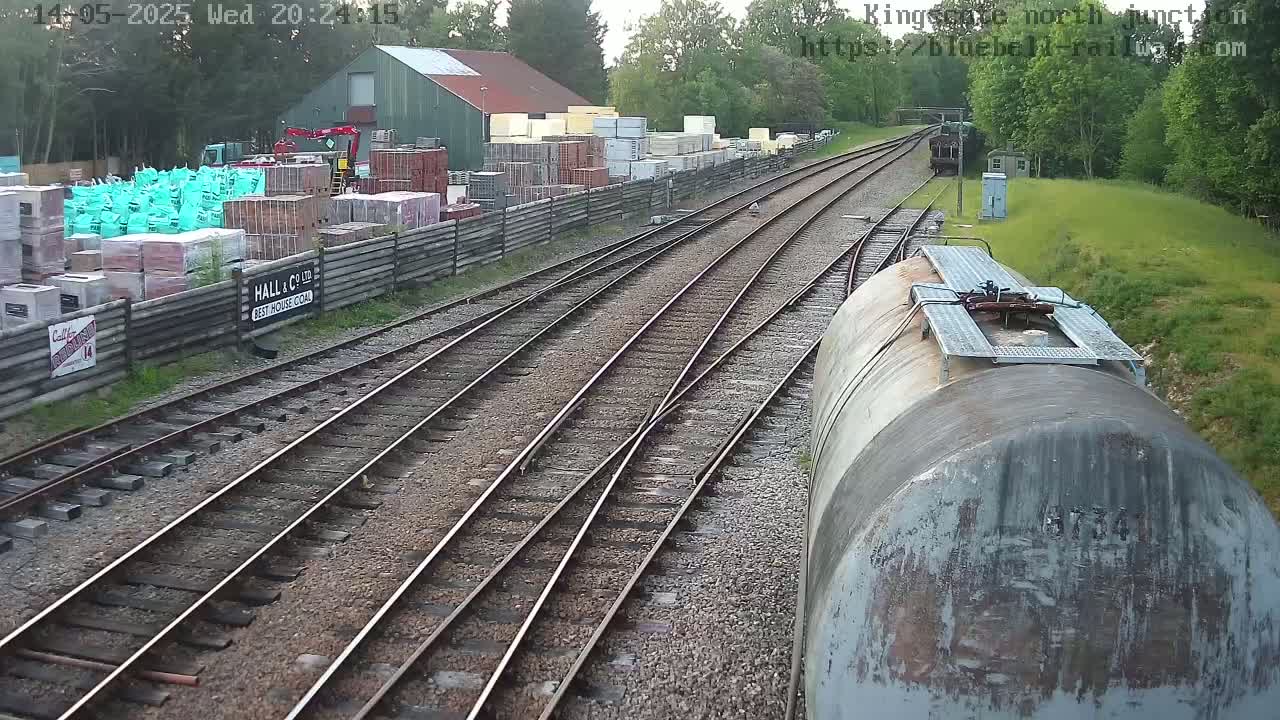 A view from a train shows multiple train tracks diverging, with a building and stacks of building materials alongside a fenced area to one side, under a partly cloudy sky.