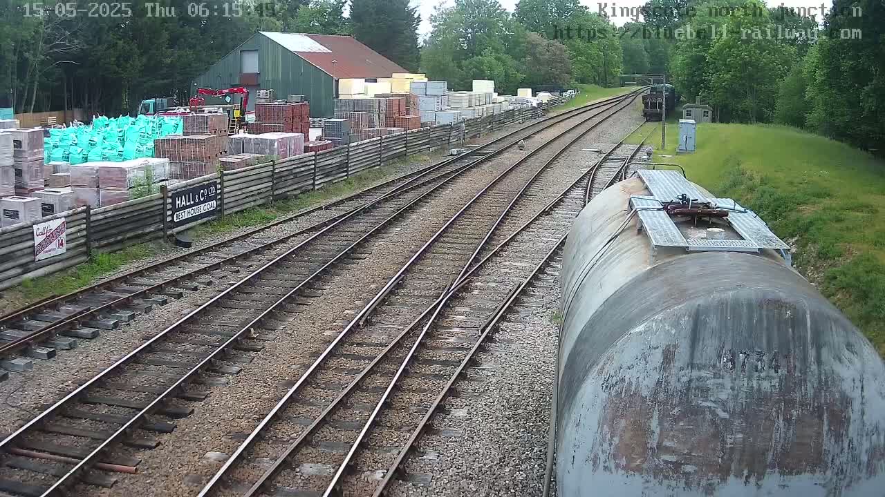 Railway tracks converge near a building and a stack of building materials, with a train car in the foreground under an overcast sky.