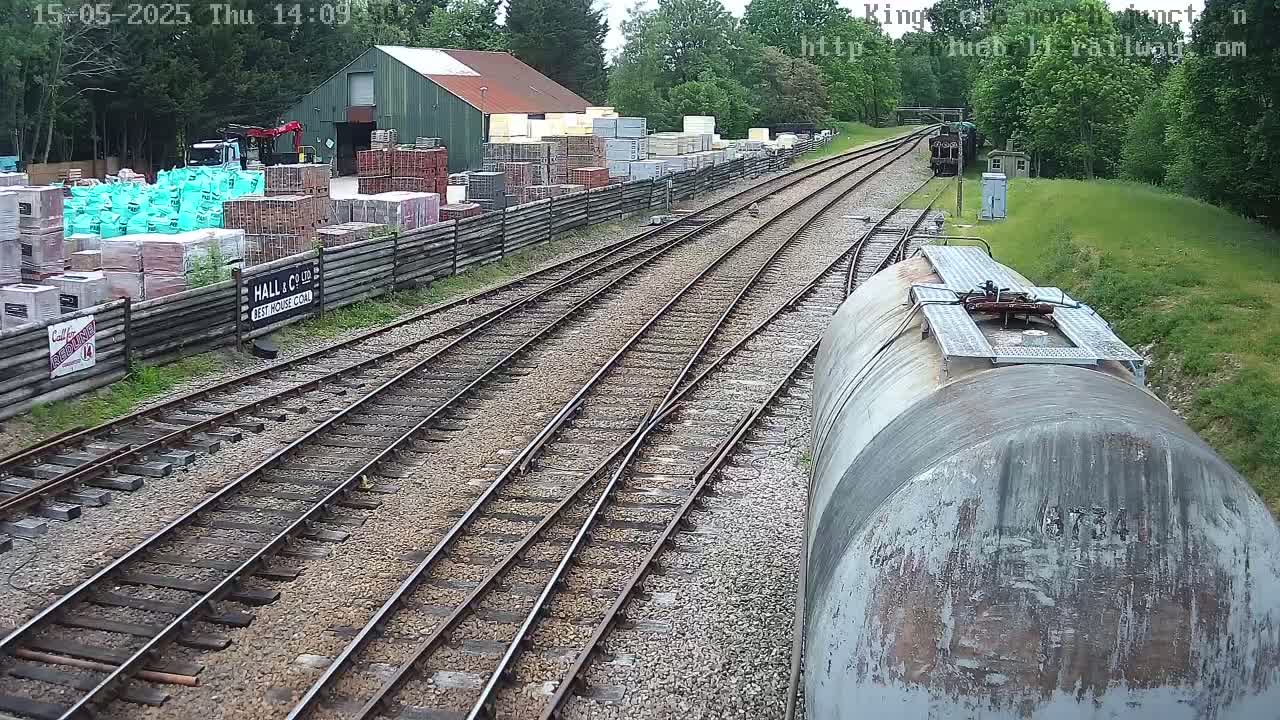 A view from a train car shows a railway track splitting into two, alongside a building and yard containing stacks of building materials, under a partly cloudy sky.