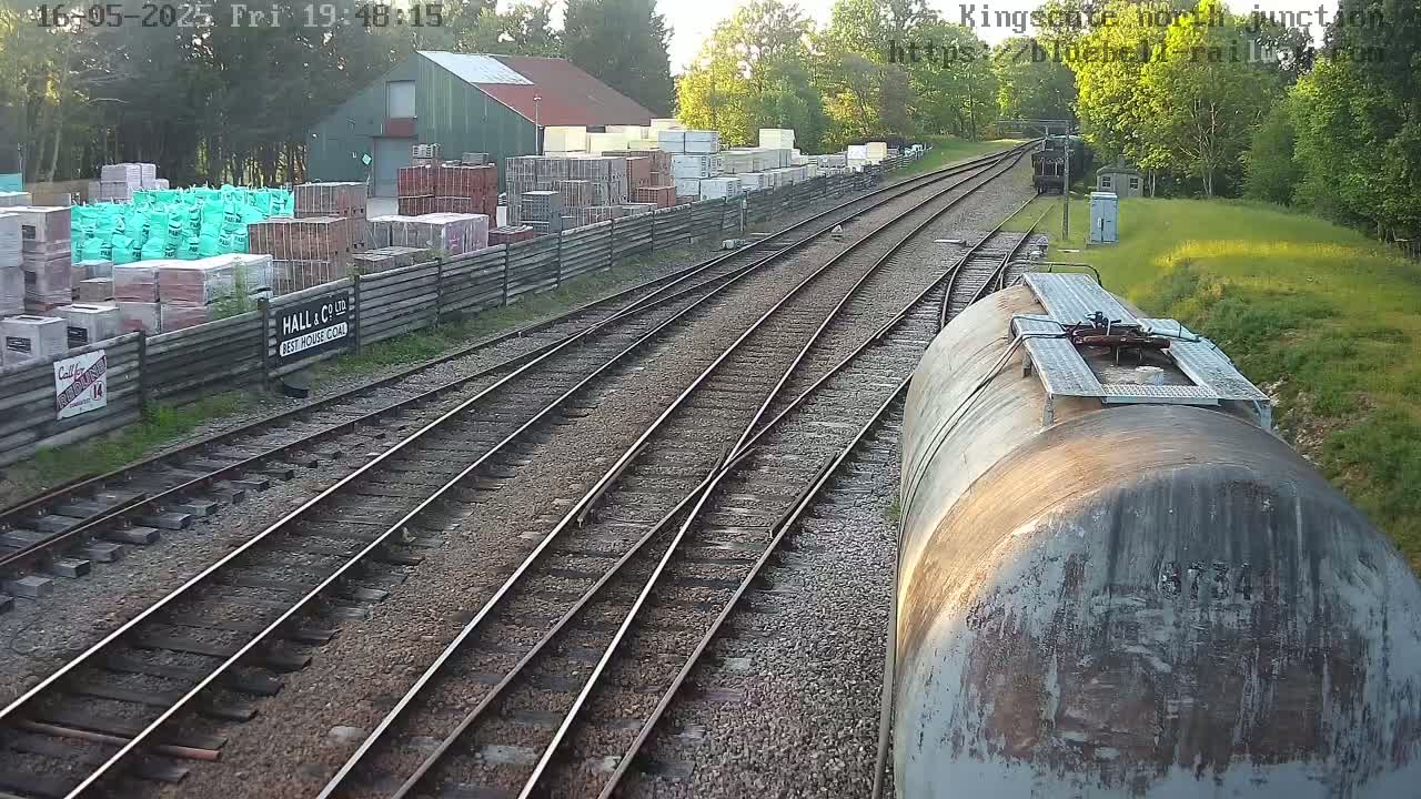 A view from a train shows multiple train tracks branching out from a central point, passing by a building with stacked building materials and a wooden fence, under partly sunny skies.