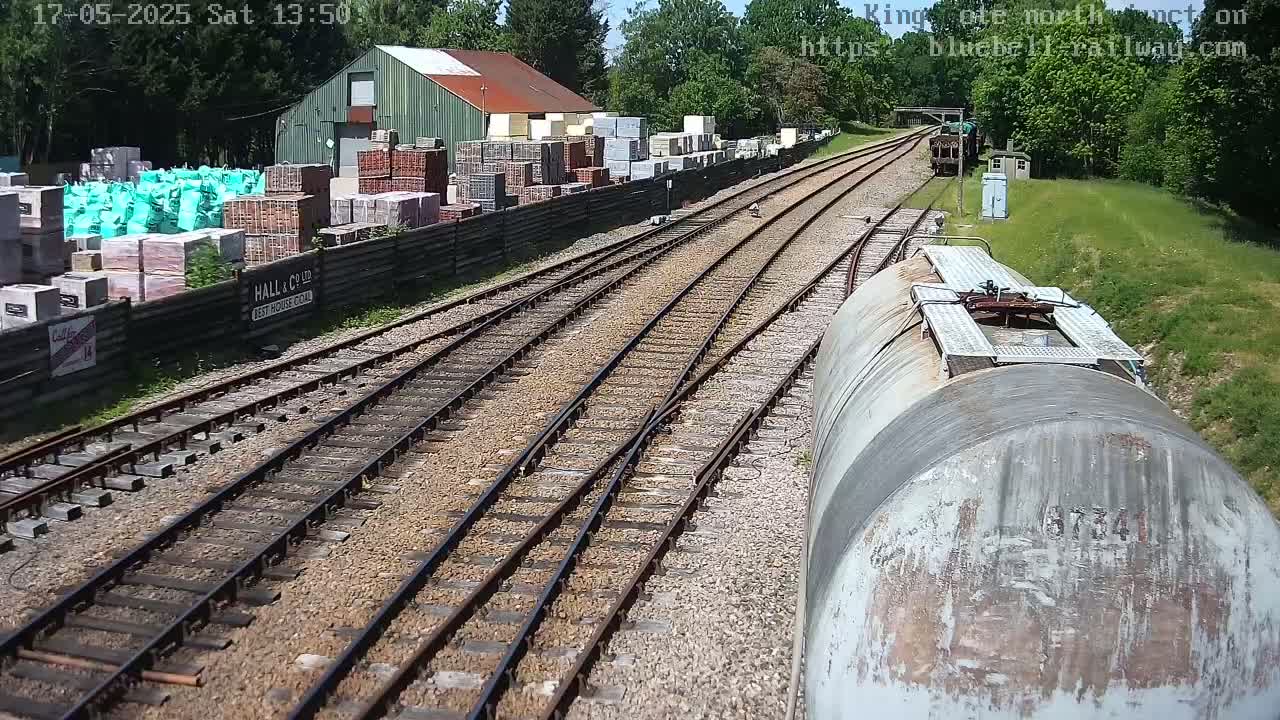 A train car’s perspective shows railway tracks splitting into multiple paths, passing by a building and a yard full of stacked building materials, all under a sunny sky.