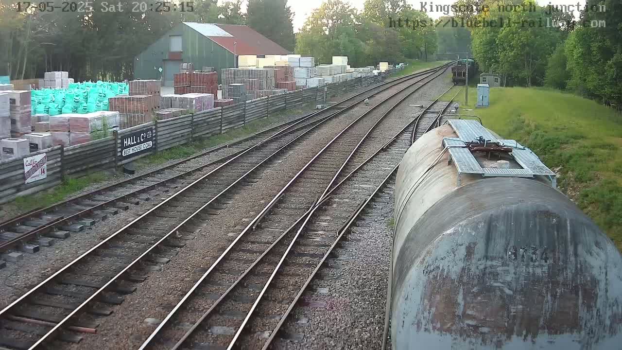 A view of railway tracks and a train car, beside a building with stacked building materials, on a partly sunny day.