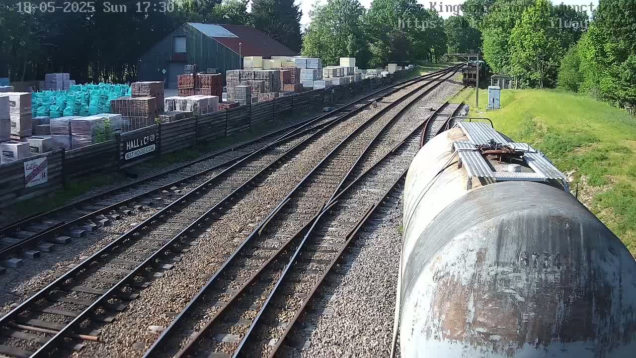 A train travels past a railway siding next to a building and a yard full of stacked building materials on a sunny day.