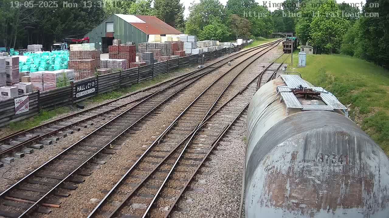 A high-angle view shows multiple train tracks branching off, with a large building containing stacks of building materials in the background on a partly sunny day.