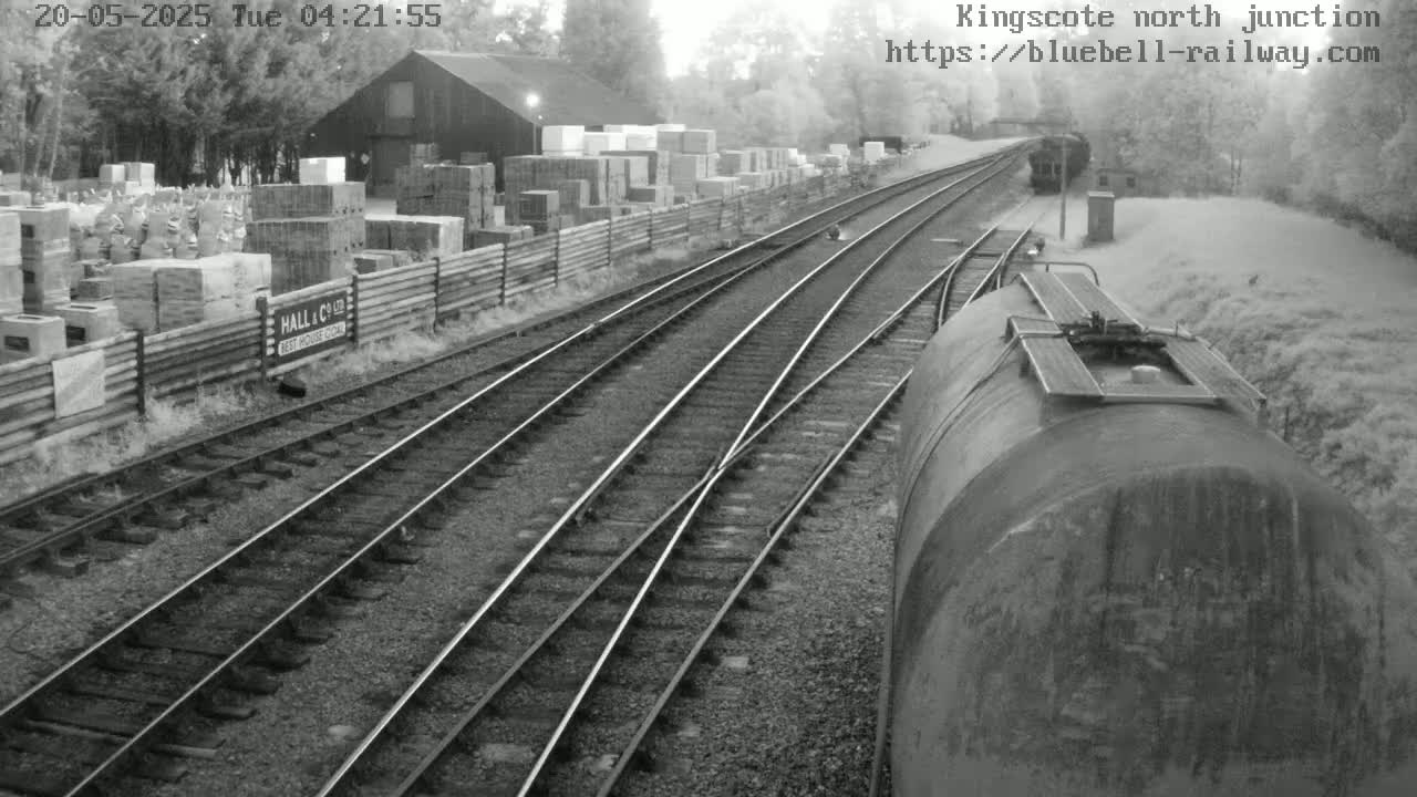 A view from a train shows multiple train tracks converging, with a building and stacks of building materials visible to the side under a hazy sky.