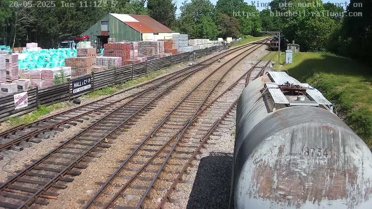 A train car travels along railway tracks past a building and a yard full of stacked building materials on a sunny day.