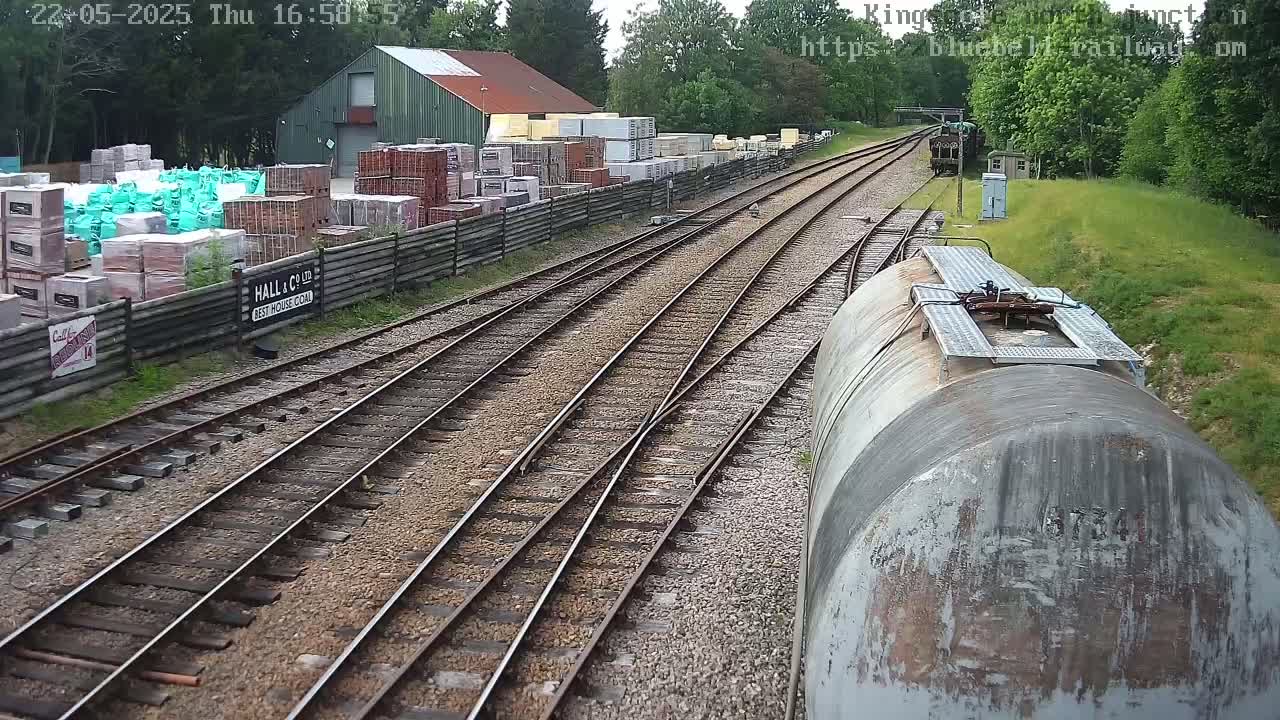 Railway tracks diverge past a building and stacks of building materials, viewed from a train car on a partly sunny day.