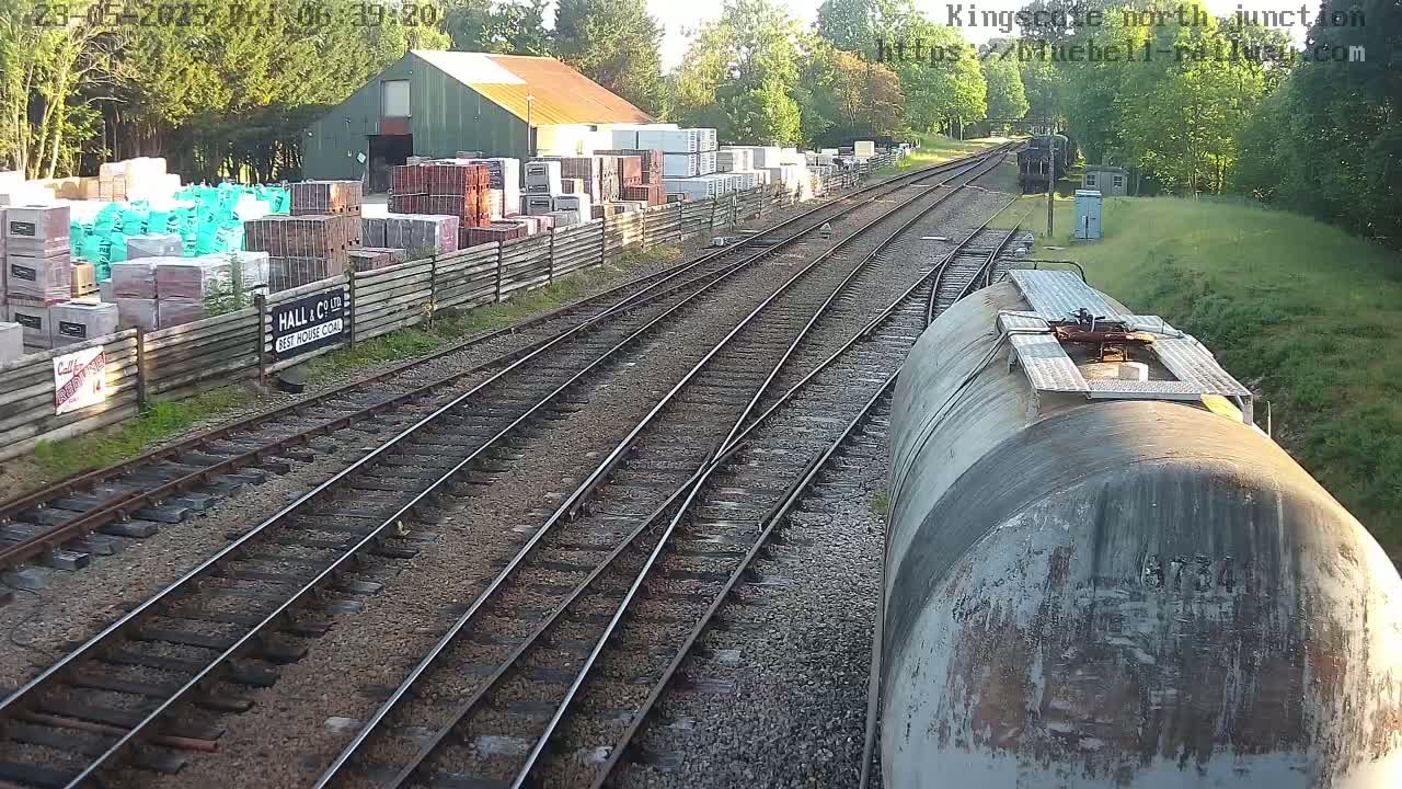A view from a train shows railway tracks converging at a junction, alongside a building with stacks of building materials and a grassy embankment under a sunny sky.